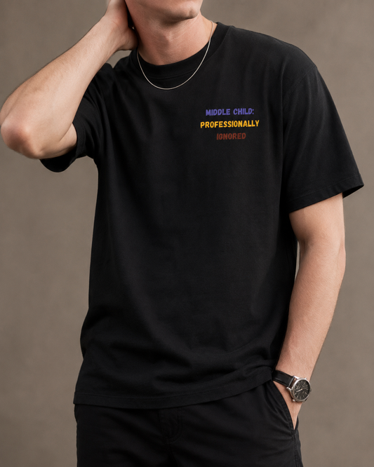 A man wearing a black crew-neck t-shirt with a small colorful graphic on the left chest that reads 'MIDDLE CHILD: PROFESSIONALLY IGNORED'. The background is a solid, warm gray studio backdrop.